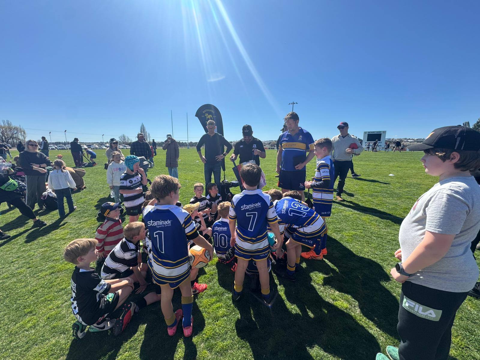 Lane Cove Minis players gathered around coaches for a sideline team talk in bright sunshine